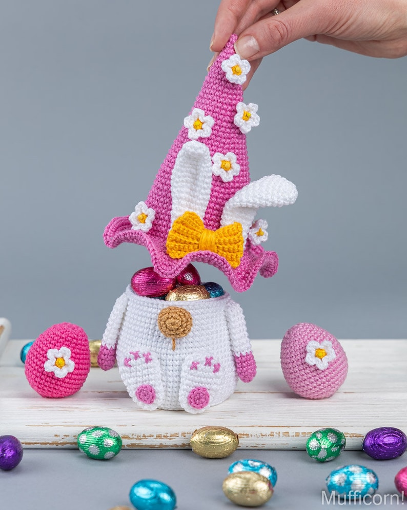 May include: A crocheted Easter bunny basket with a pink hat and a yellow bow. The basket is filled with chocolate eggs and there are more eggs scattered around it.