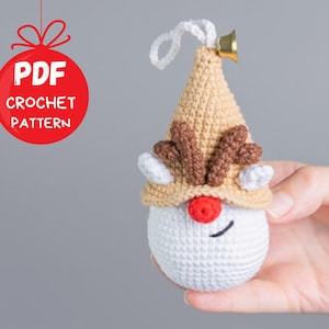 May include: Crochet pattern for a reindeer ornament. The ornament is white with a brown hat and antlers. It has a red nose and a black stitched smile. The ornament is held in a hand.
