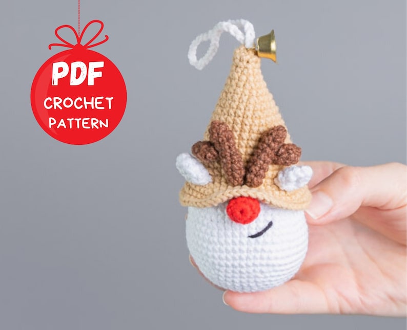 May include: Crochet pattern for a reindeer ornament. The ornament is white with a brown hat and antlers. It has a red nose and a black stitched smile. The ornament is held in a hand.