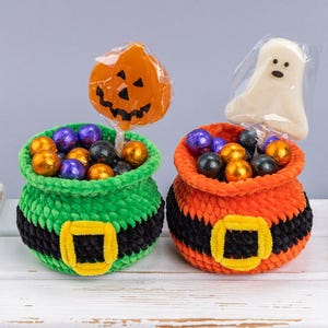 May include: Two crocheted pots, one green and one orange, each featuring a black band with a yellow buckle. The green pot holds a pumpkin-shaped lollipop and purple and orange candies. The orange pot holds a ghost-shaped lollipop and candies.