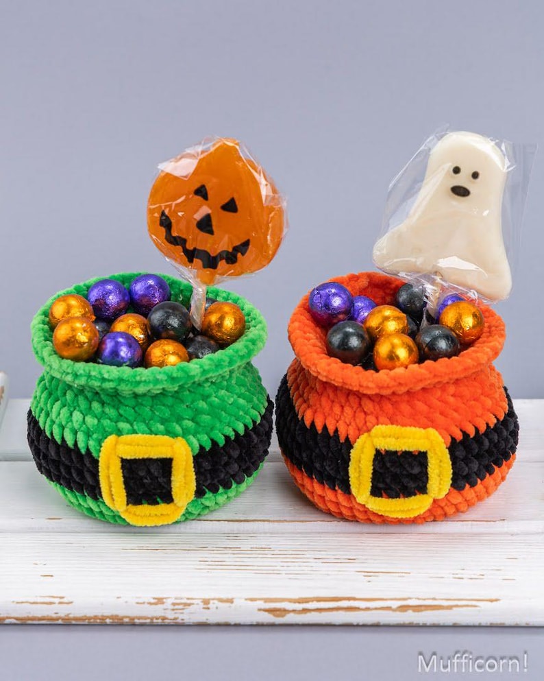 May include: Two crocheted pots, one green and one orange, each featuring a black band with a yellow buckle. The green pot holds a pumpkin-shaped lollipop and purple and orange candies. The orange pot holds a ghost-shaped lollipop and candies.