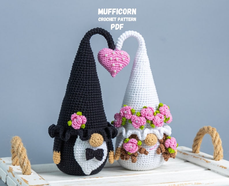 May include: Two crocheted gnomes, one black and one white, holding a pink heart. They are wearing flower crowns and the black gnome has a white bow tie. The gnomes are sitting on a white tray with rope handles. The text "MUFFICORN CROCHET PATTERN PDF" is at the top of the image.