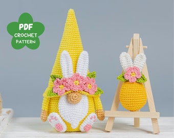 Puede incluir: Un gnomo de Pascua de crochet con un sombrero amarillo, orejas de conejo blancas y flores rosas. El gnomo sostiene una barba blanca y tiene pies rosas. El gnomo está sentado sobre una superficie blanca. Un pequeño huevo de Pascua de crochet con orejas de conejo y flores rosas está en un caballete de madera.