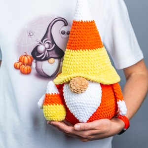 May include: A crocheted candy corn gnome with a white beard and orange and yellow hat. The gnome is holding a smaller candy corn gnome in its hand.