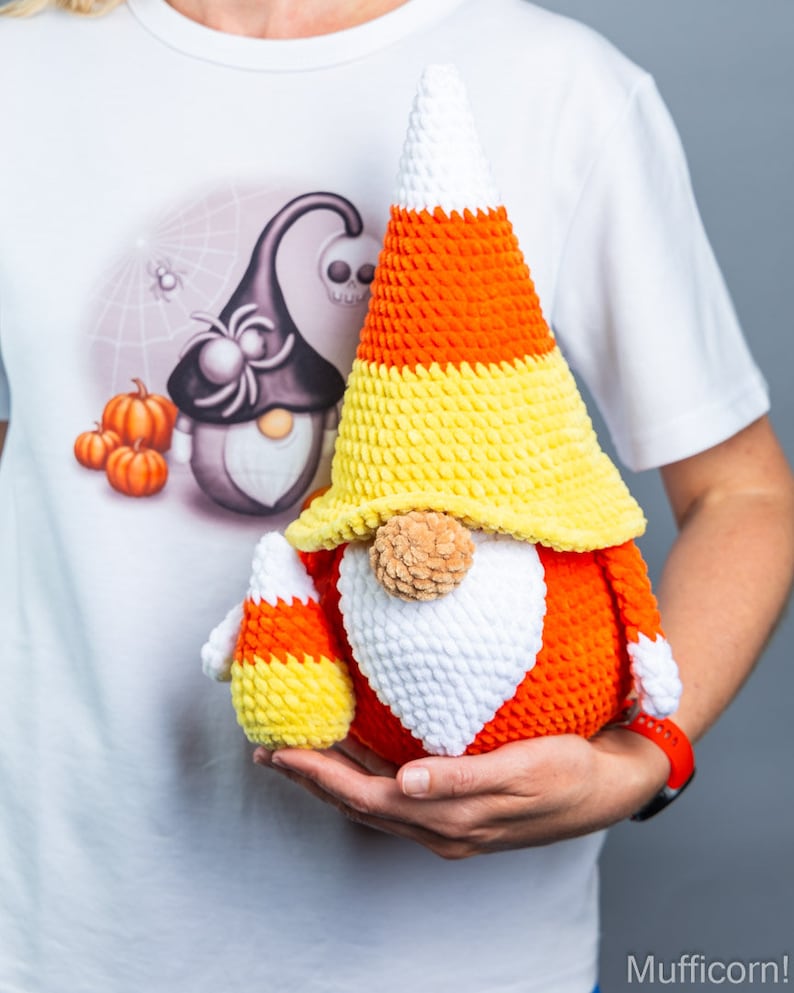 May include: A crocheted candy corn gnome with a white beard and orange and yellow hat. The gnome is holding a smaller candy corn gnome in its hand.