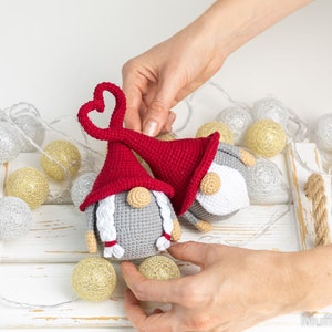 May include: Two crocheted gnomes with red and gray hats. The gnome on the left has a heart-shaped hat and white braids. The gnome on the right has a red hat with a white pom-pom. The gnomes are sitting on a white tray with string lights.