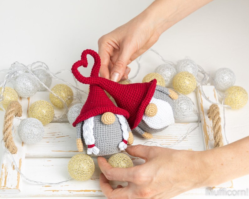 May include: Two crocheted gnomes with red and gray hats. The gnome on the left has a heart-shaped hat and white braids. The gnome on the right has a red hat with a white pom-pom. The gnomes are sitting on a white tray with string lights.
