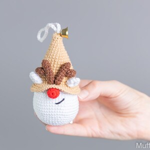 May include: A crocheted reindeer ornament with a brown hat, white face, red nose, and brown antlers. The ornament has a gold bell attached to the top.