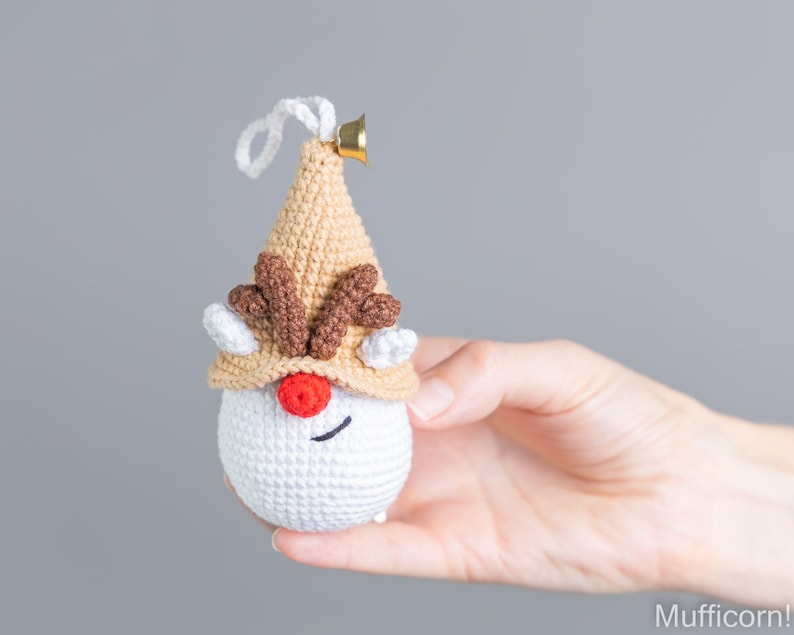 May include: A crocheted reindeer ornament with a brown hat, white face, red nose, and brown antlers. The ornament has a gold bell attached to the top.