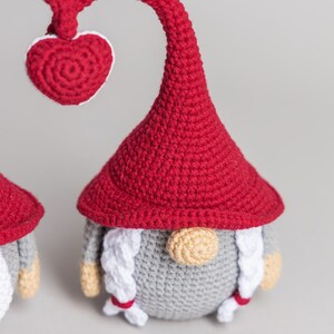 May include: A red crocheted gnome with a heart-shaped hat and a white beard. The gnome is standing on a gray surface.