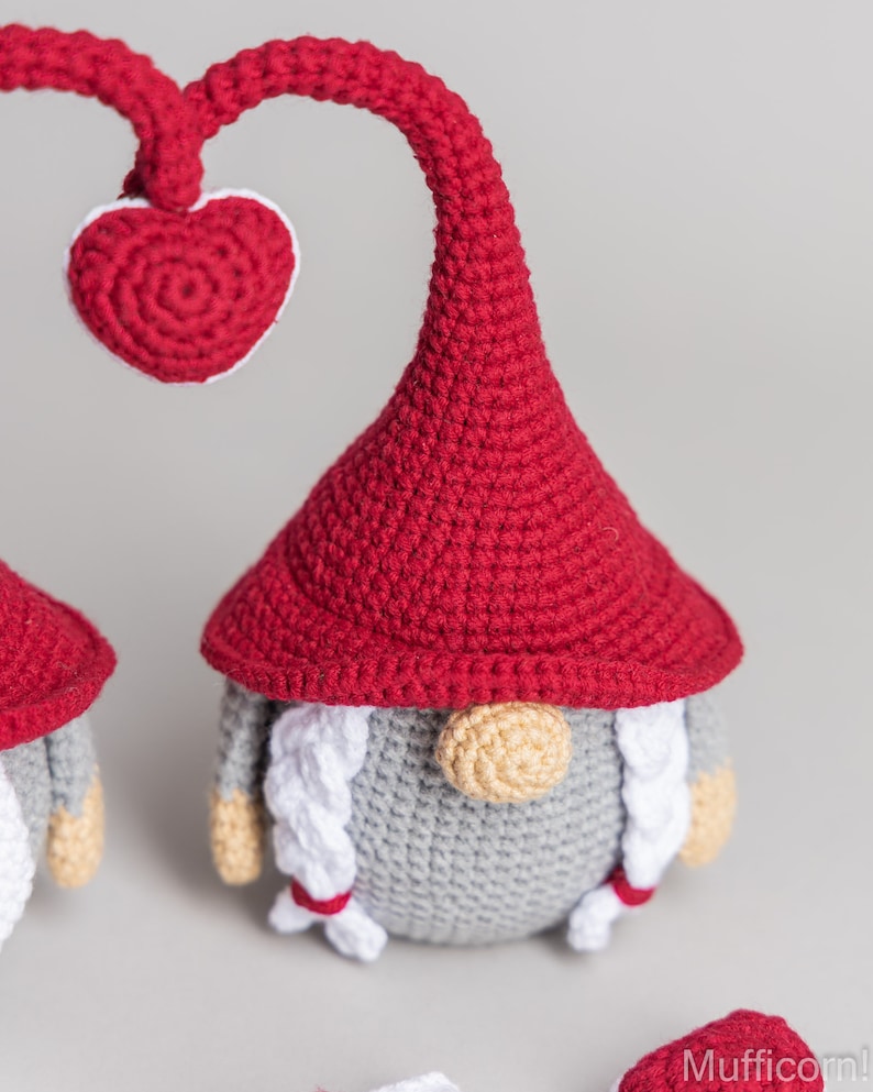 May include: A red crocheted gnome with a heart-shaped hat and a white beard. The gnome is standing on a gray surface.
