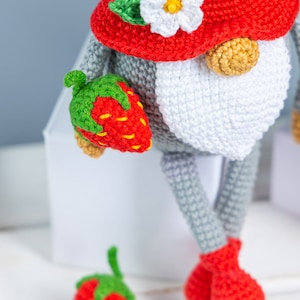 Op de afbeelding: Een gehaakte gnoompup met een rode muts met een witte bloem en groene bladeren. De gnoom houdt een rode gehaakte aardbei in zijn hand. Er ligt nog een aardbei op het witte oppervlak onder de gnoom.