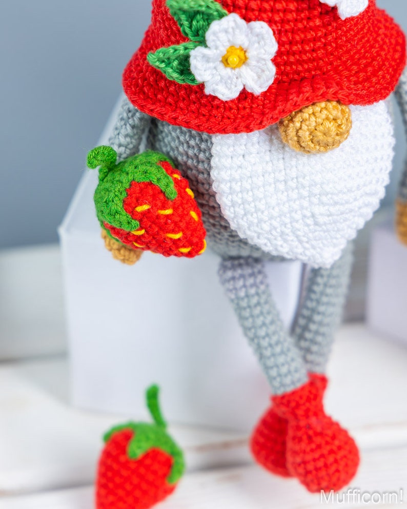 Op de afbeelding: Een gehaakte gnoompup met een rode muts met een witte bloem en groene bladeren. De gnoom houdt een rode gehaakte aardbei in zijn hand. Er ligt nog een aardbei op het witte oppervlak onder de gnoom.