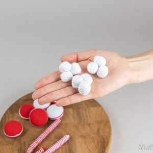 Puede incluir: Una tabla de servir de madera muestra varios art&iacute;culos de ganchillo hechos a mano. Se sostienen bolas de ganchillo blancas en una mano, mientras que caramelos y macarons de ganchillo rojos y blancos est&aacute;n dispuestos en la tabla. La imagen tiene un fondo neutro.