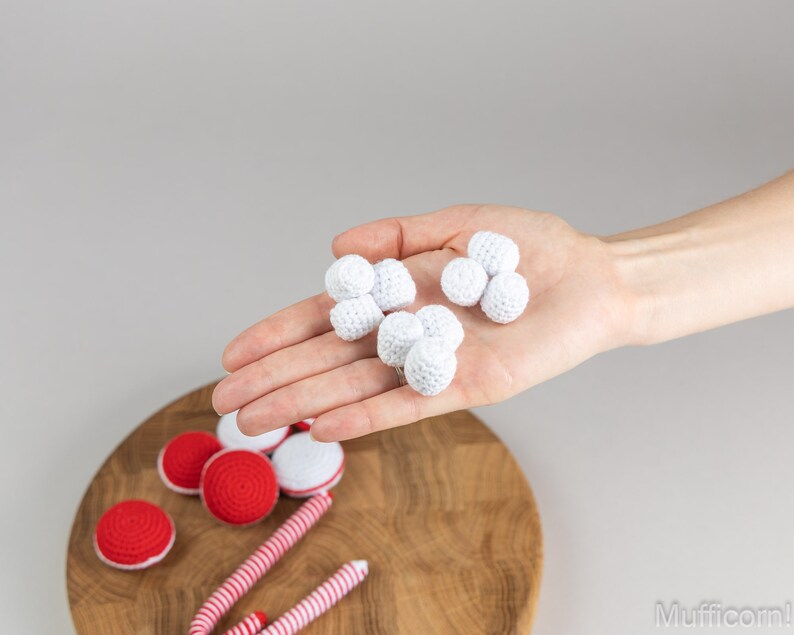 Puede incluir: Una tabla de servir de madera muestra varios art&iacute;culos de ganchillo hechos a mano. Se sostienen bolas de ganchillo blancas en una mano, mientras que caramelos y macarons de ganchillo rojos y blancos est&aacute;n dispuestos en la tabla. La imagen tiene un fondo neutro.