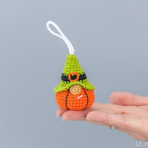 May include: A crocheted pumpkin gnome ornament with a green hat, black belt, and orange pumpkin body. The gnome has a white beard and a smiling face.