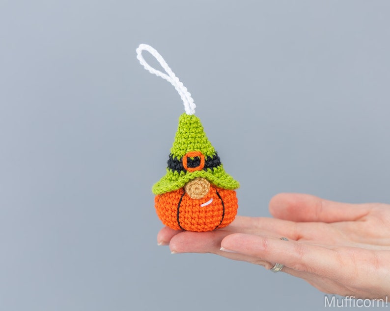 May include: A crocheted pumpkin gnome ornament with a green hat, black belt, and orange pumpkin body. The gnome has a white beard and a smiling face.