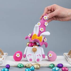 May include: A crocheted Easter bunny basket with a pink hat and white flowers. The basket is filled with colorful chocolate eggs and is sitting on a white tray with other Easter eggs.