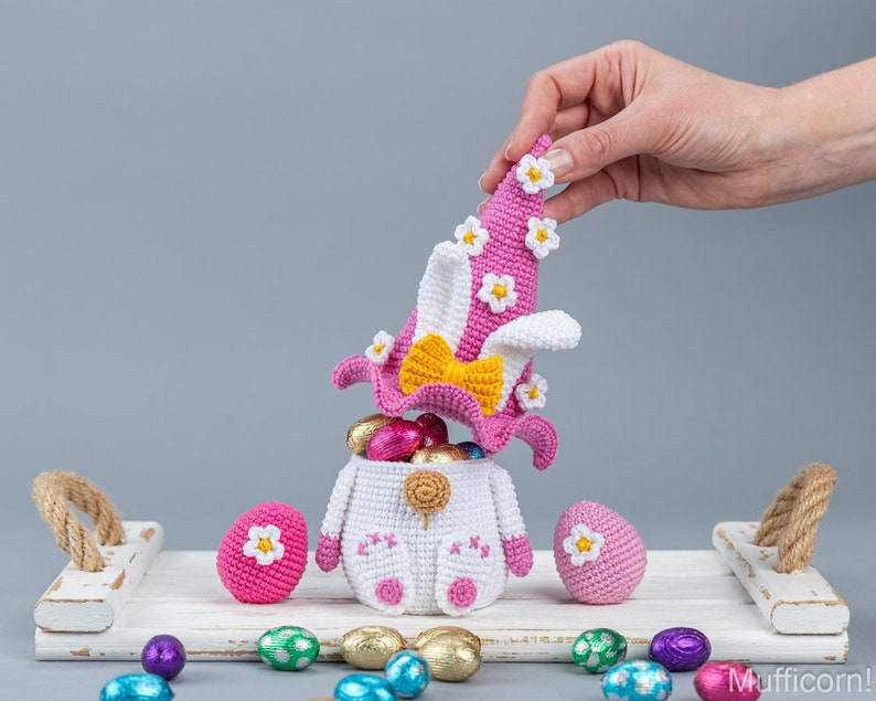 May include: A crocheted Easter bunny basket with a pink hat and white flowers. The basket is filled with colorful chocolate eggs and is sitting on a white tray with other Easter eggs.