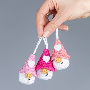 May include: Three handmade crochet gnomes with white bodies, pink hats, and white heart accents. Each gnome has a small, tan nose and a white string for hanging. The gnomes are held against a gray background.