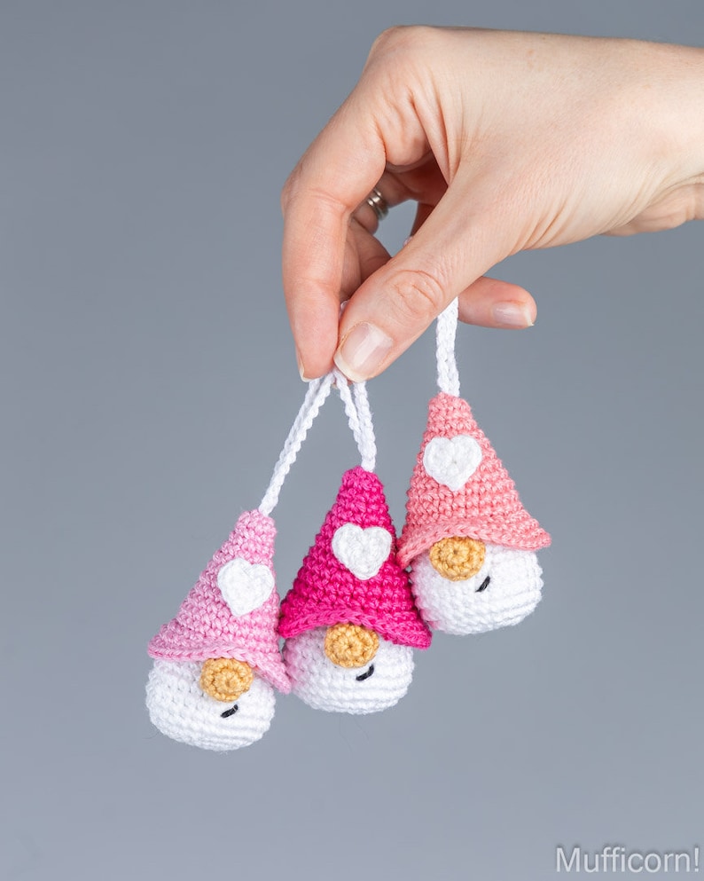 May include: Three handmade crochet gnomes with white bodies, pink hats, and white heart accents. Each gnome has a small, tan nose and a white string for hanging. The gnomes are held against a gray background.