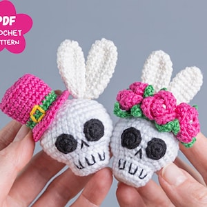 Op de afbeelding: Twee witte gehaakte schedel ornamenten, een met een roze hoge hoed met een groene band en de andere met een roze bloemenkrans. De tekst PDF Crochet Pattern is zichtbaar in de linkerbovenhoek van de afbeelding.