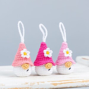 May include: Three handmade crochet gnome ornaments. Each gnome has a white body, a pink or fuchsia hat with a white and yellow flower, and a small tan nose. White string loops are attached for hanging.