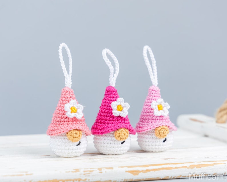 May include: Three handmade crochet gnome ornaments. Each gnome has a white body, a pink or fuchsia hat with a white and yellow flower, and a small tan nose. White string loops are attached for hanging.