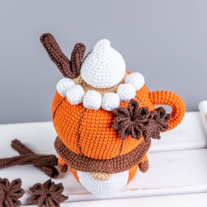 Puede incluir: Una taza naranja de ganchillo con un asa marr&oacute;n, cubierta con crema batida blanca, astas marrones y malvaviscos blancos. Una bufanda marr&oacute;n envuelve la taza. Acentos decorativos marrones en forma de estrella y palitos de canela est&aacute;n dispersos alrededor de la taza.