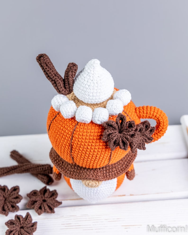 Puede incluir: Una taza naranja de ganchillo con un asa marr&oacute;n, cubierta con crema batida blanca, astas marrones y malvaviscos blancos. Una bufanda marr&oacute;n envuelve la taza. Acentos decorativos marrones en forma de estrella y palitos de canela est&aacute;n dispersos alrededor de la taza.