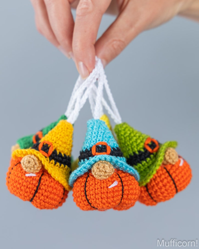 May include: Four crocheted gnome ornaments with pumpkin-shaped bodies, each with a different colored hat: green, yellow, blue, and green. The gnomes have white beards and black belts.