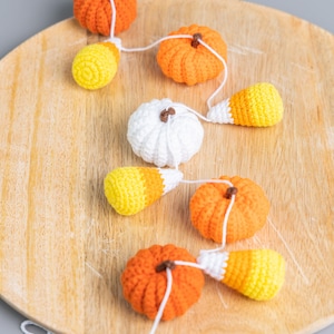 May include: A crocheted garland featuring orange pumpkins, white pumpkins, and yellow and orange candy corn. The garland is strung on a white cord and is displayed on a wooden surface.