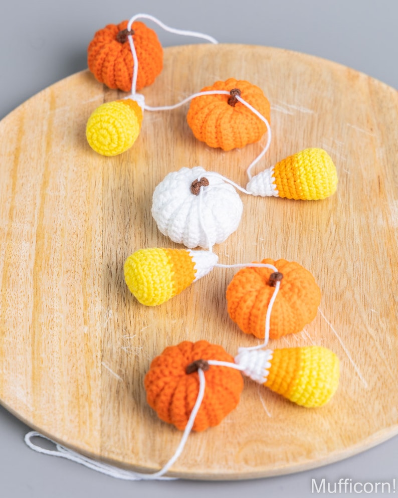 May include: A crocheted garland featuring orange pumpkins, white pumpkins, and yellow and orange candy corn. The garland is strung on a white cord and is displayed on a wooden surface.
