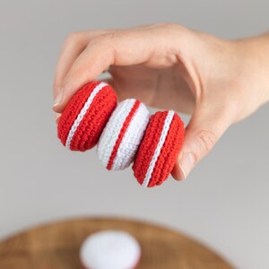 Puede incluir: Una mano sostiene tres macarons de ganchillo. Dos son rojos con una franja blanca, y uno es blanco con una franja roja. Los macarons est&aacute;n hechos a mano. El fondo es gris claro, con una superficie de madera debajo.