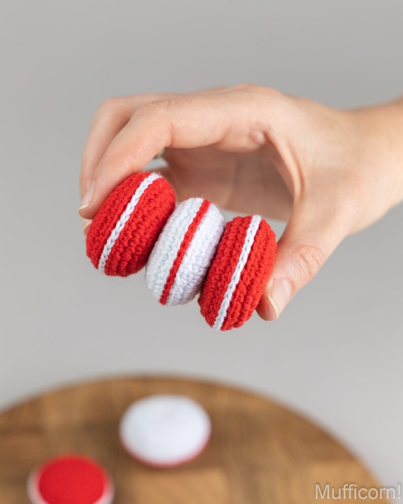 Puede incluir: Una mano sostiene tres macarons de ganchillo. Dos son rojos con una franja blanca, y uno es blanco con una franja roja. Los macarons est&aacute;n hechos a mano. El fondo es gris claro, con una superficie de madera debajo.