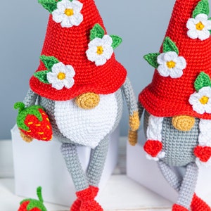 Op de afbeelding: Twee gehaakte gnomen met rode mutsen en witte bloemen. De gnomen zitten op witte oppervlakken en hebben grijze lichamen en witte baarden. E&eacute;n gnome houdt een rode aardbei vast.