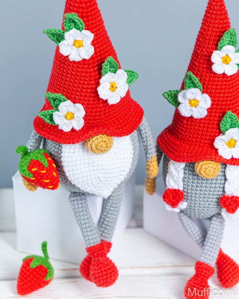 Op de afbeelding: Twee gehaakte gnomen met rode mutsen en witte bloemen. De gnomen zitten op witte oppervlakken en hebben grijze lichamen en witte baarden. E&eacute;n gnome houdt een rode aardbei vast.