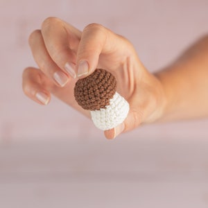 May include: A hand holding a small crocheted mushroom toy. The mushroom is brown on top and white on the bottom.