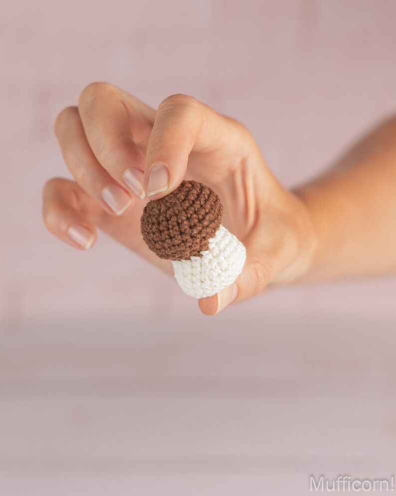 May include: A hand holding a small crocheted mushroom toy. The mushroom is brown on top and white on the bottom.