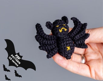 May include: A black crocheted bat with yellow embroidered eyes and a black star on its chest. The bat is being held in a hand.