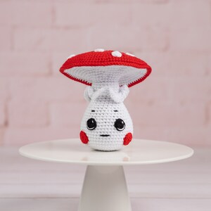 May include: A white crocheted mushroom with a red and white spotted cap. The mushroom has a cute face with black eyes and a small mouth. It is sitting on a white cake stand.