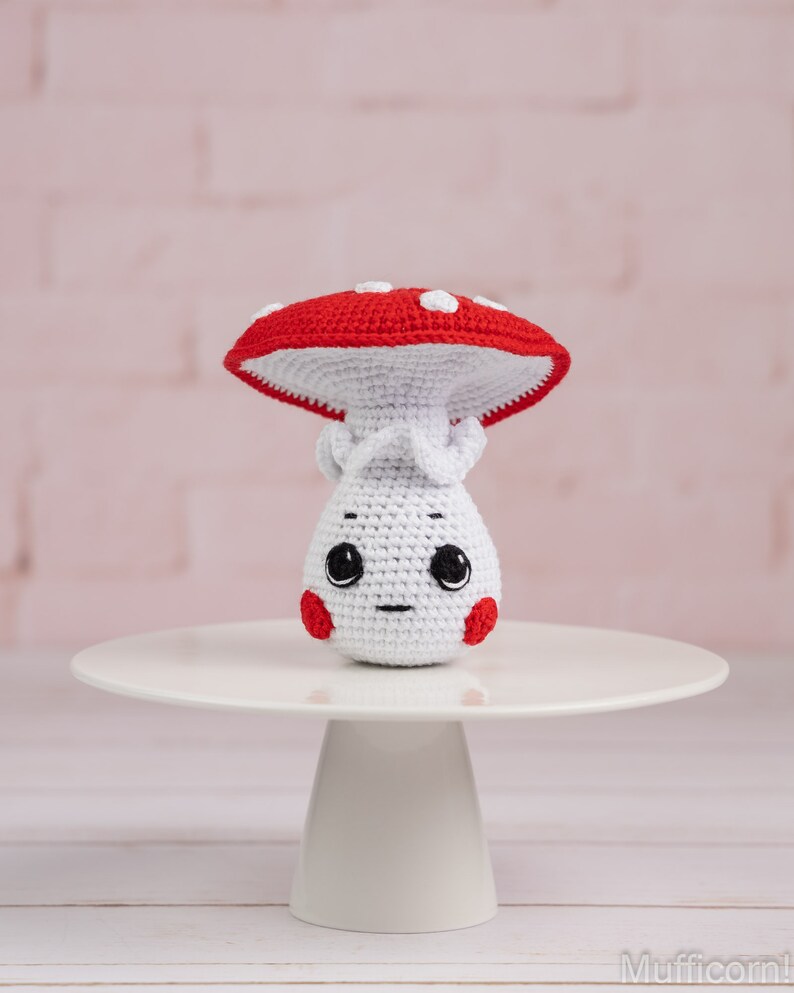 May include: A white crocheted mushroom with a red and white spotted cap. The mushroom has a cute face with black eyes and a small mouth. It is sitting on a white cake stand.