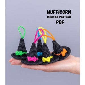 Può includere: Una mano mostra diversi piccoli cappelli da strega neri all'uncinetto, ognuno con un fiocco e un anello colorato. I cappelli presentano dettagli verdi, rosa, gialli, blu e arancioni. L'immagine include il testo "MUFFICORN CROCHET PATTERN PDF."