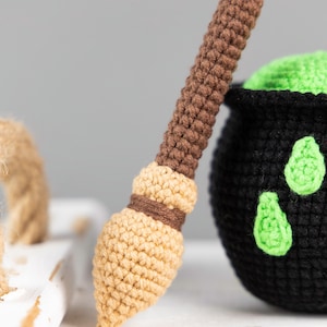 May include: A brown and tan crocheted broomstick with a green cauldron in the background. The cauldron is black with green accents.
