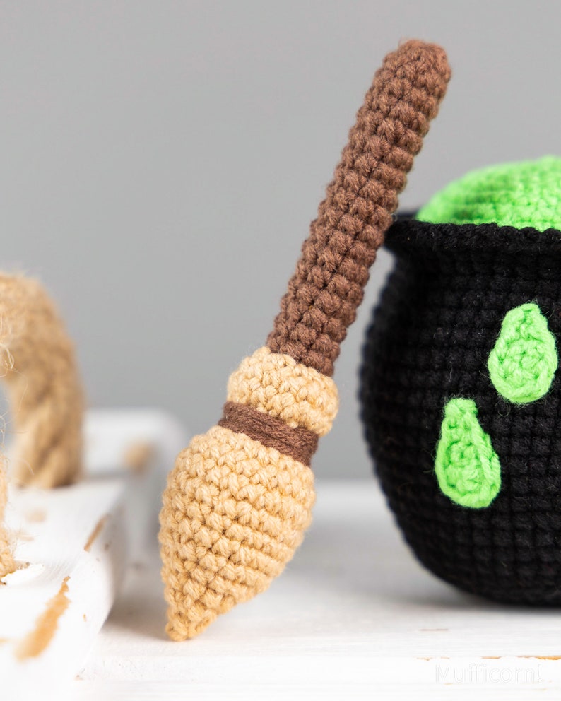May include: A brown and tan crocheted broomstick with a green cauldron in the background. The cauldron is black with green accents.