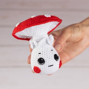 May include: A crocheted mushroom toy with a red cap and white body. The mushroom has a cute face with black eyes and a small smile. The toy is held in a hand.
