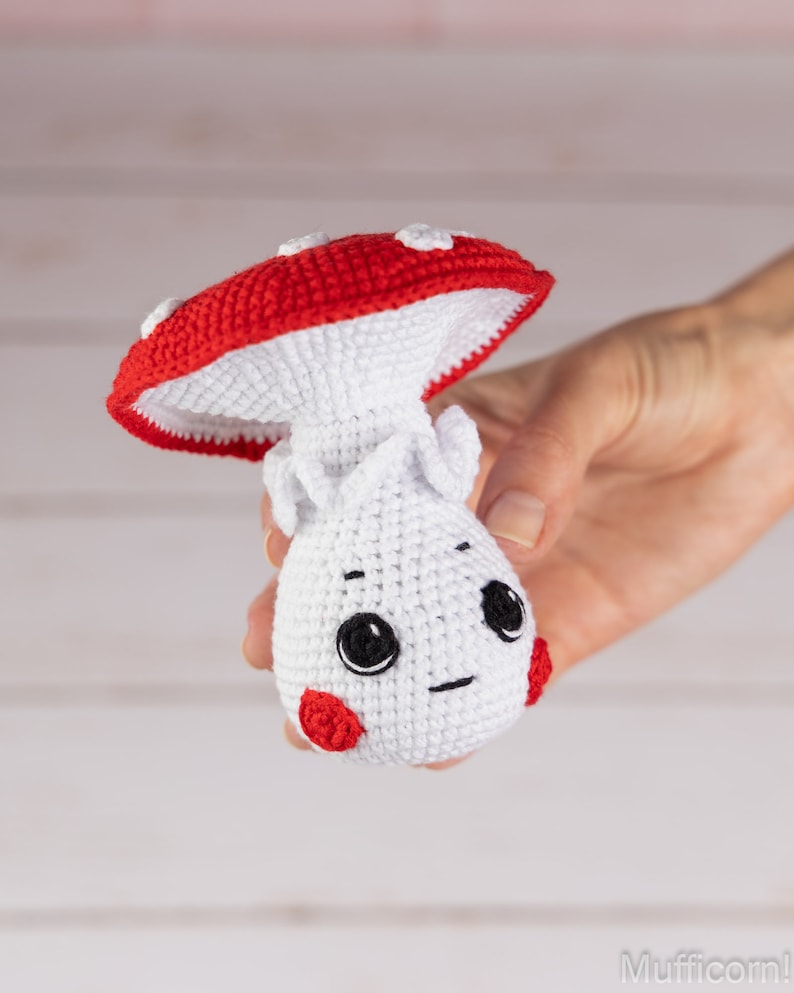 May include: A crocheted mushroom toy with a red cap and white body. The mushroom has a cute face with black eyes and a small smile. The toy is held in a hand.