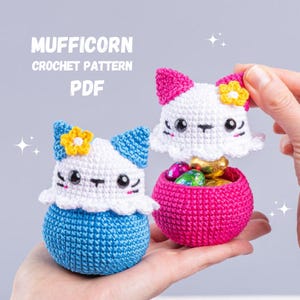 Può includere: Due contenitori a forma di gatto all'uncinetto, uno blu e uno rosa, ciascuno con una testa bianca, un fiore giallo e dettagli del viso neri. Il contenitore rosa è aperto, rivelando uova di cioccolato. Il testo "MUFFICORN CROCHET PATTERN PDF" è in alto.