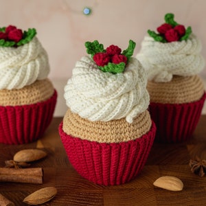 May include: Three crocheted cupcakes with red and white frosting and green holly leaves. The cupcakes are sitting on a wooden surface with cinnamon sticks and star anise.