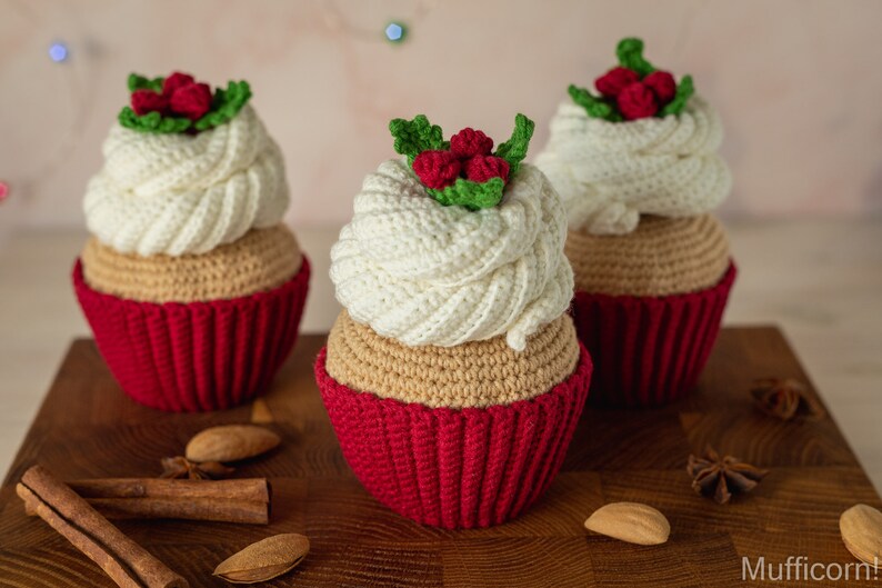 May include: Three crocheted cupcakes with red and white frosting and green holly leaves. The cupcakes are sitting on a wooden surface with cinnamon sticks and star anise.